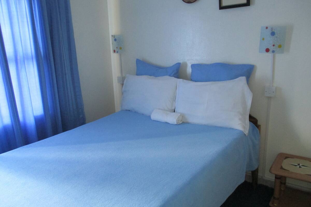 A single bed with a blue bedspread and white pillows in a room with a blue curtain on one side.