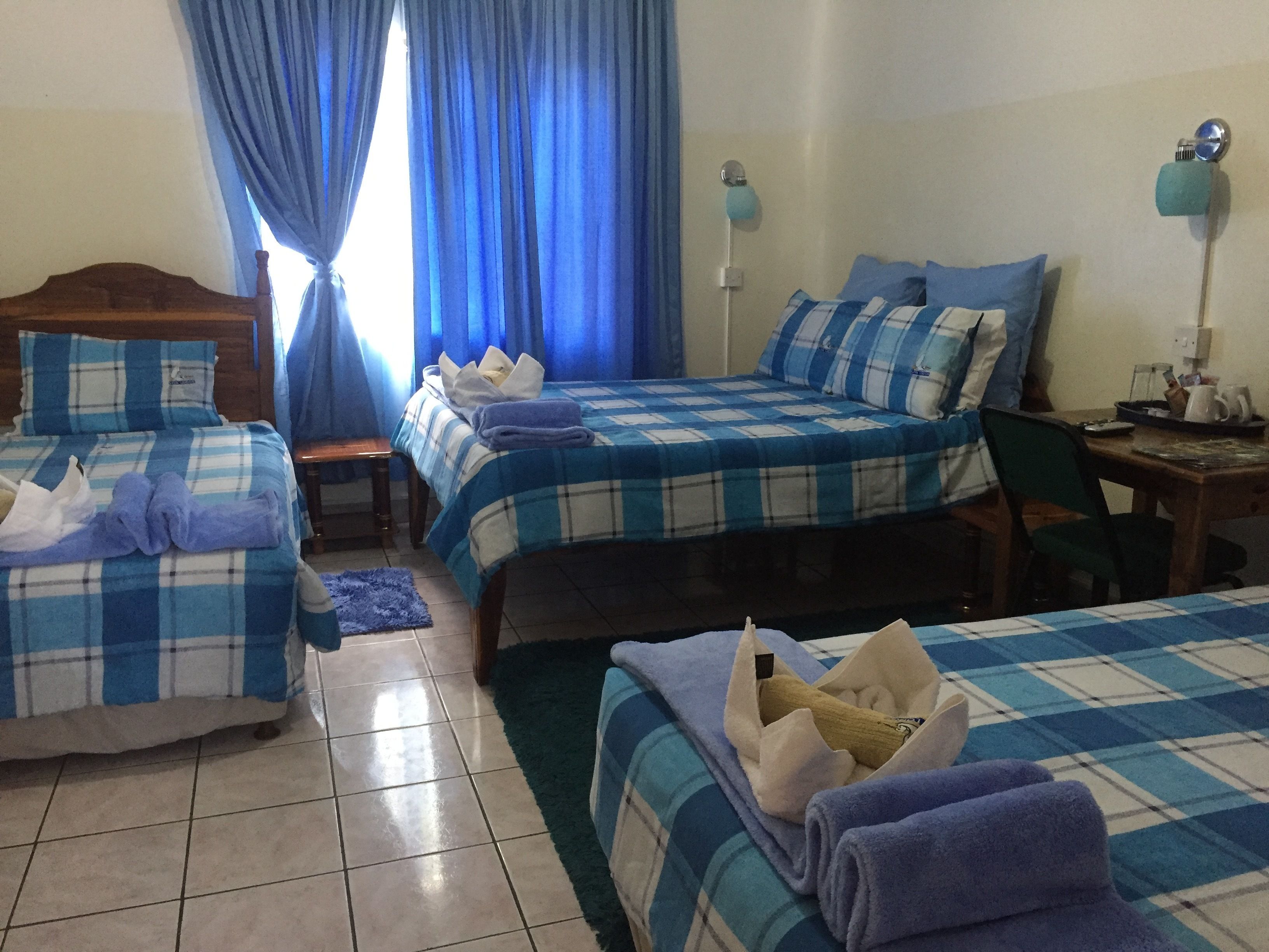 Habitación de hotel con tres camas, cada una con ropa de cama azul y blanca a cuadros. Se observa un cabecero de madera, una mesita y una silla. El piso es de baldosas y hay una ventana con cortinas azules.