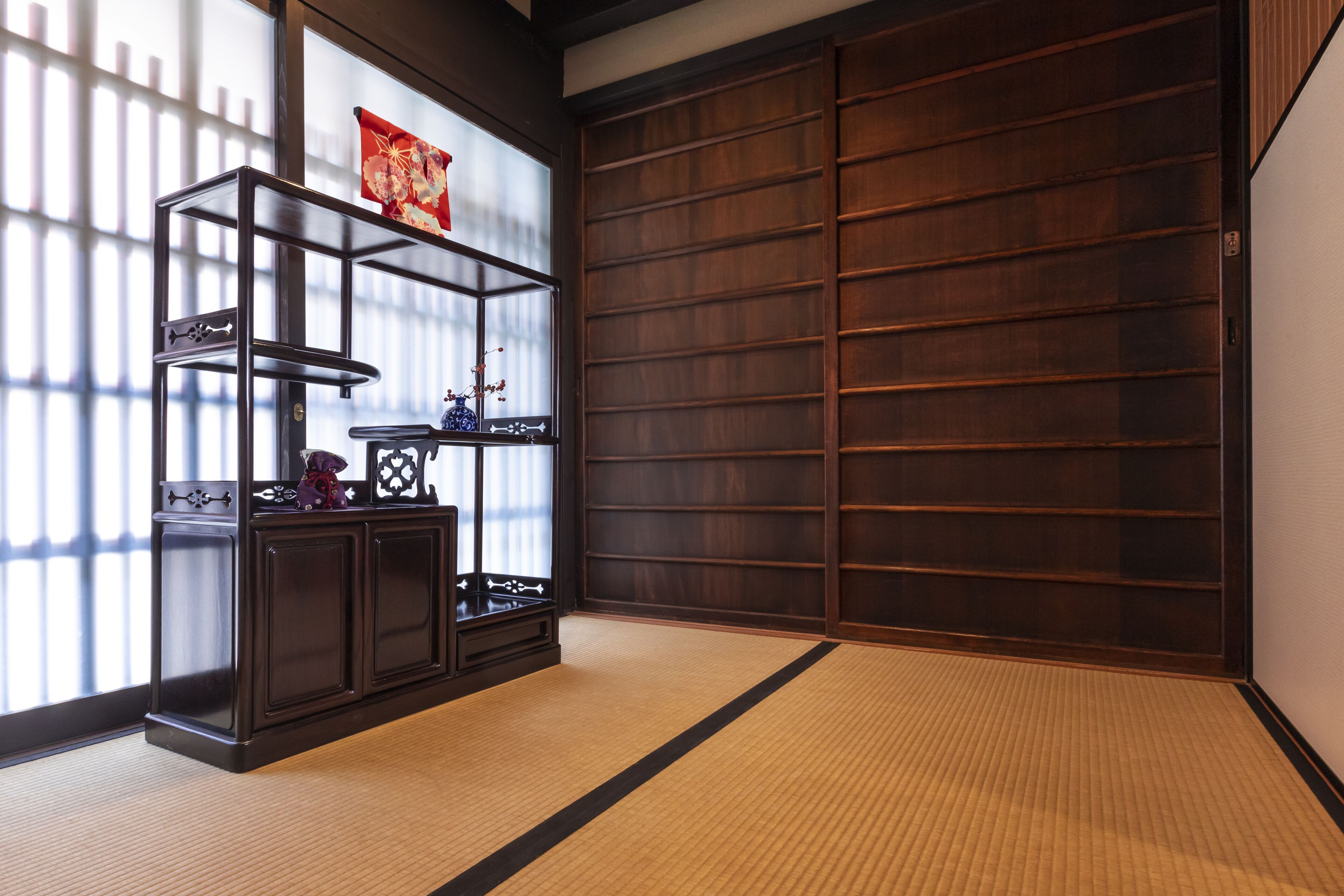Ein traditioneller japanischer Raum mit Tatami-Böden, einem dunklen Holzregal mit Dekoration und einer Schiebetür.