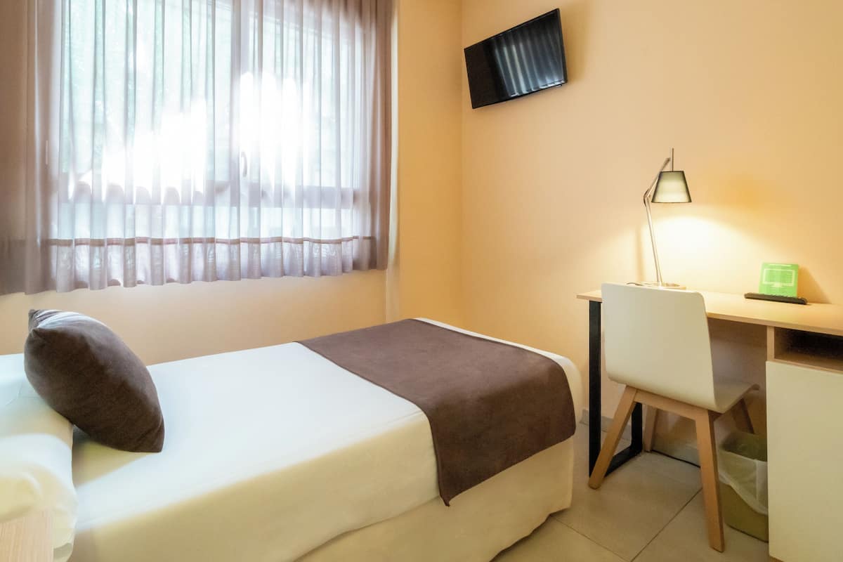 Un dormitorio con cama, escritorio, silla, lámpara y televisor en la pared.