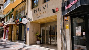 Entrada de un hotel con puerta de vidrio, un cartel de Nuvi Hotels y una estanquera cerca.