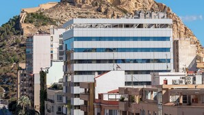 Un edificio moderno con tejado plano y múltiples ventanas, ubicado en una colina con un afloramiento rocoso al fondo.