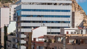 Un edificio moderno con techo plano y varios balcones, situado en una ciudad con una colina destacada al fondo.
