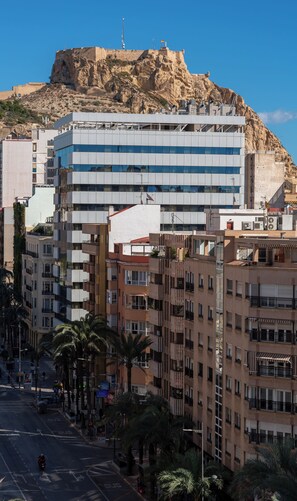 Un edificio moderno con techo plano y varios balcones, situado en una ciudad con una colina destacada al fondo.