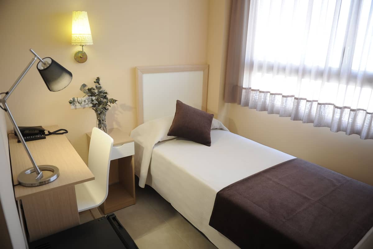 Habitación de hotel con cama, escritorio, silla, lámpara y ventana con cortinas.