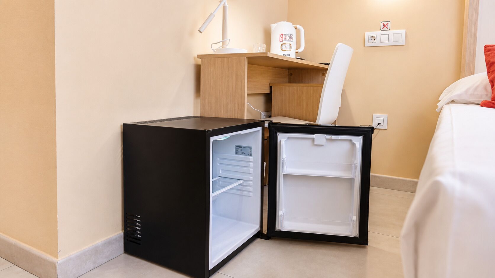 Un refrigerador compacto negro con compartimento congelador, ubicado en una habitación de hotel con un escritorio de madera, una silla y una cama con ropa blanca.