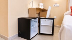 Un refrigerador compacto negro con compartimento congelador, ubicado en una habitación de hotel con un escritorio de madera, una silla y una cama con ropa blanca.
