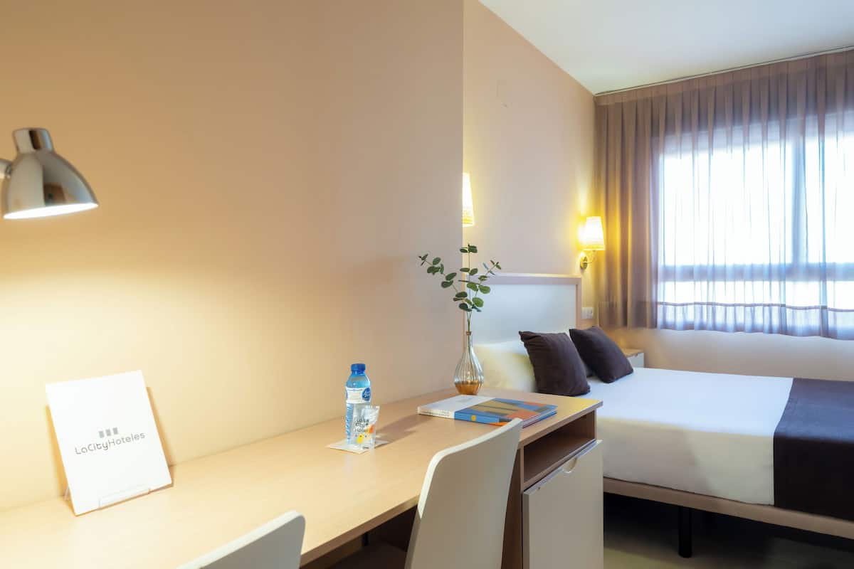 Habitaci ón de hotel con cama, escritorio, silla, lámpara, botella de agua y un cartel de LaCityHotels.