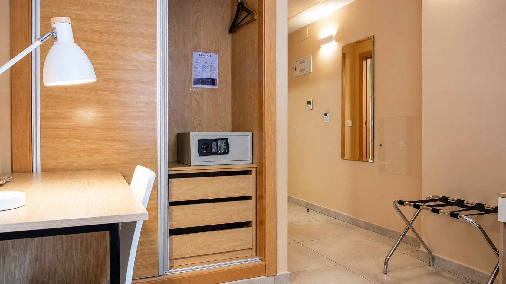Habitación de hotel con escritorio de madera, lámpara de escritorio blanca, microondas y carrito para equipaje negro.