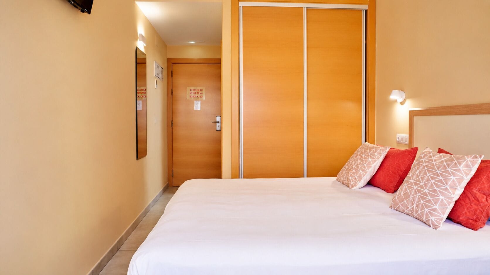 Una habitación de hotel con una cama, almohadas, un armario de madera y una lámpara fijada a la pared.