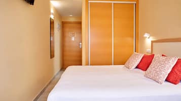 Una habitación de hotel con una cama, almohadas, un armario de madera y una lámpara fijada a la pared.