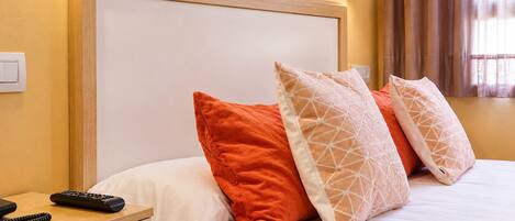 A hotel room with a bed, pillows, a bedside table, a remote control, and a wall-mounted light fixture.