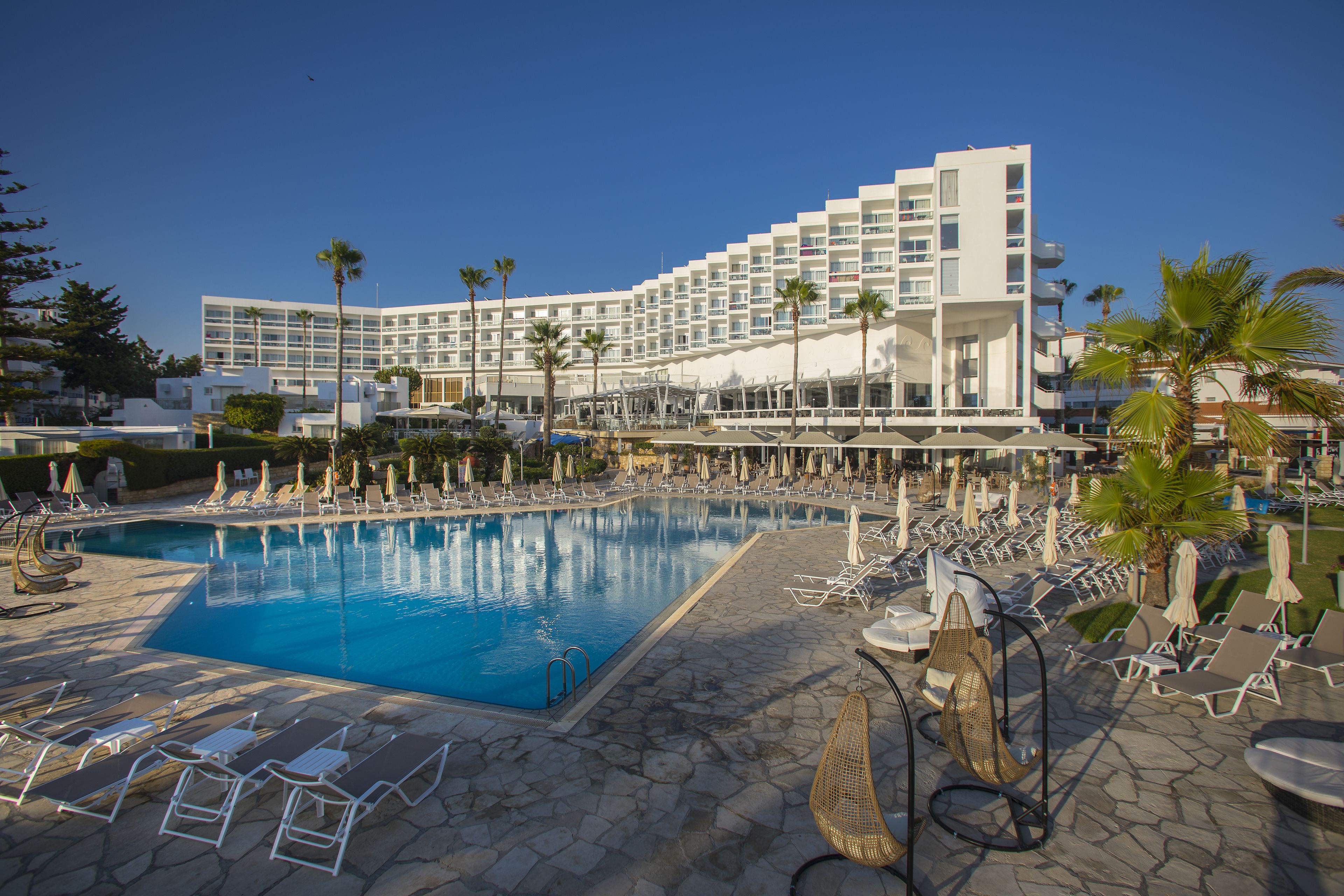 A hotel with a large pool, sun loungers, and palm trees.