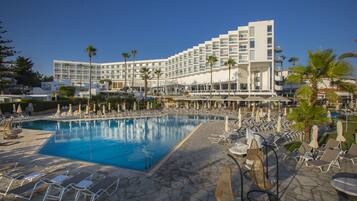 A hotel with a large pool, sun loungers, and palm trees.
