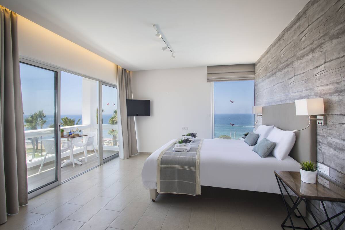Een moderne hotelkamer met een groot bed, een balkon met een tafel en stoelen, een televisie en uitzicht op de zee.
