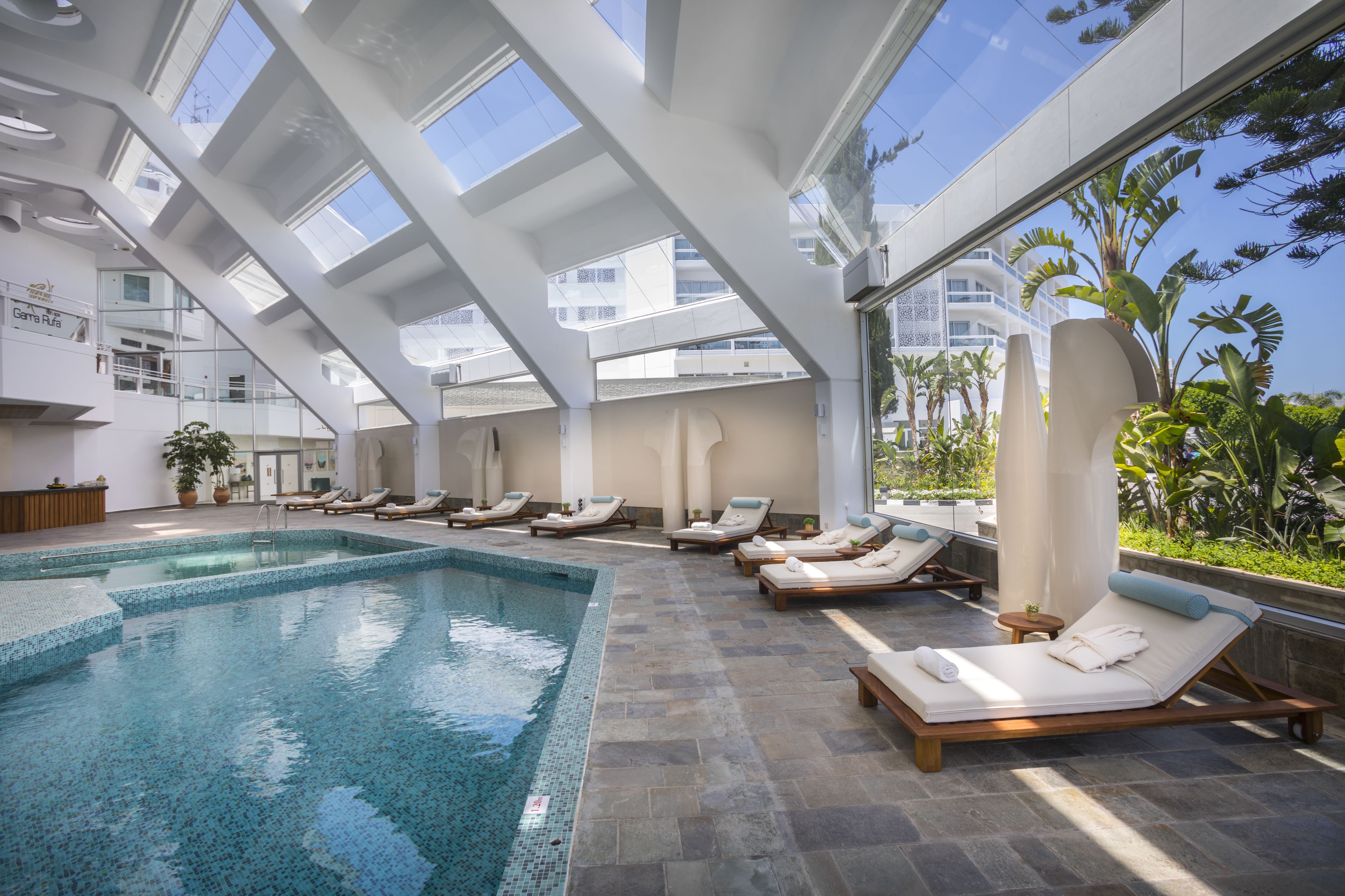 A modern indoor swimming pool with lounge chairs and a glass ceiling.