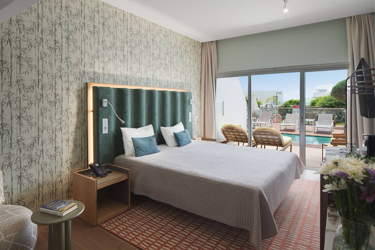 Een hotelkamer met een bed, bureau, stoel, klein tafeltje, nachtkastje, raam en uitzicht op een zwembad en terras.