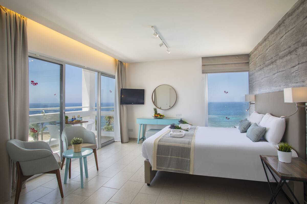 Een moderne hotelkamer met een groot bed, een balkon met uitzicht op zee, een bureau met stoel en een aan de muur bevestigde televisie.