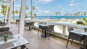 A terrace with tables and chairs, overlooking a pool and beach with palm trees. - Leonardo Plaza Cypria Maris Beach Hotel & Spa (Geroskipou)