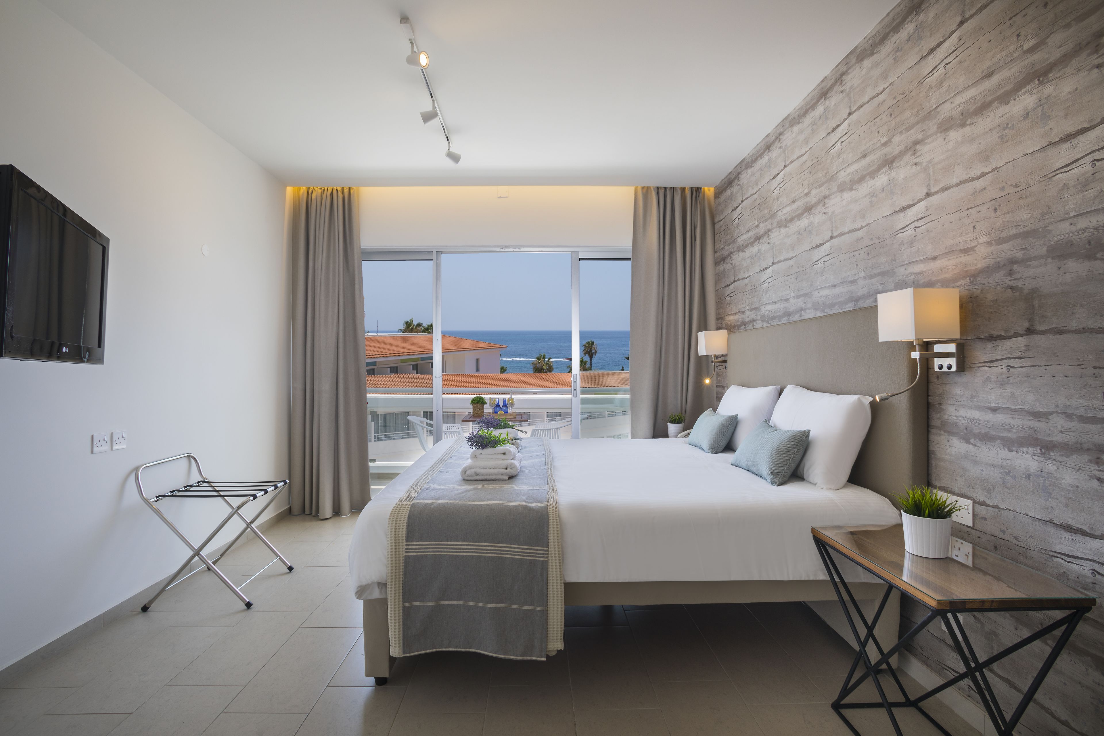 Una habitación de hotel moderna con una cama grande, un panel de madera decorativo, vista al océano, una pequeña mesa con una planta y un televisor montado en la pared.