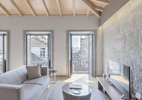 Uma sala de estar moderna com sofá, mesa de centro e televisão. Há janelas amplas e uma varanda com vista para edifícios.