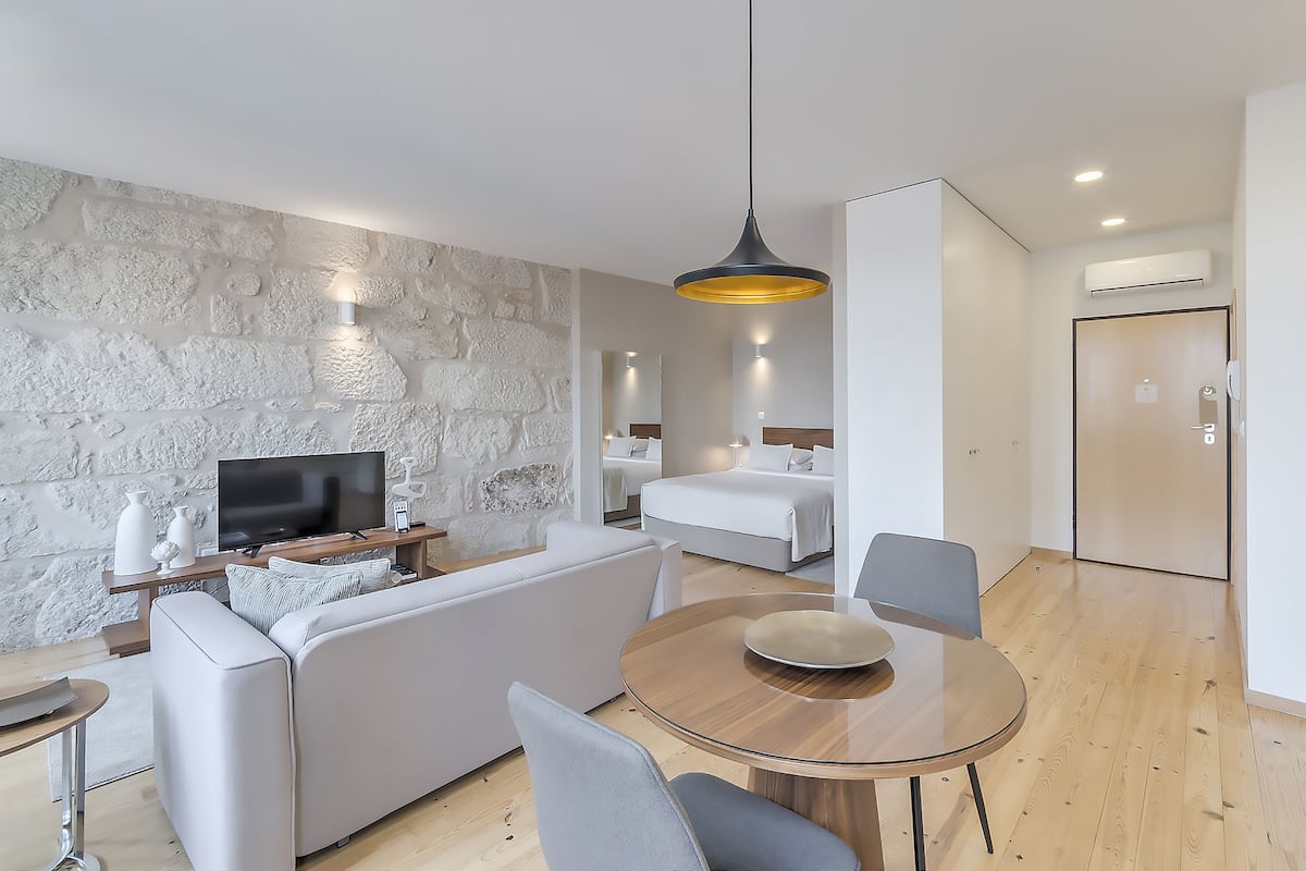 A modern hotel room with a stone accent wall, a flat-screen TV, a sofa, a dining table, and a bed with a headboard.