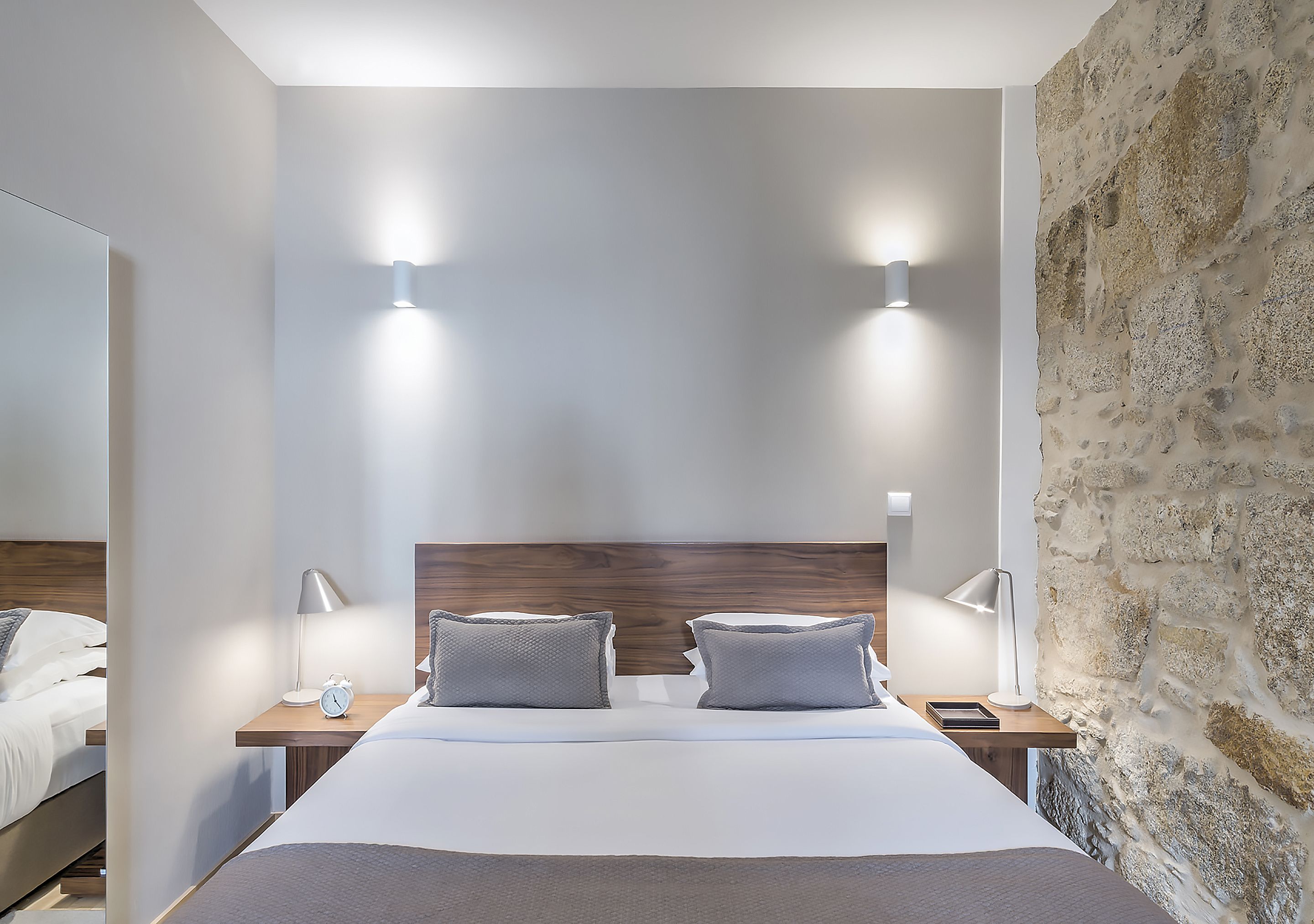 A modern hotel room with a large bed, two bedside tables with lamps, a wooden headboard, and a stone wall.
