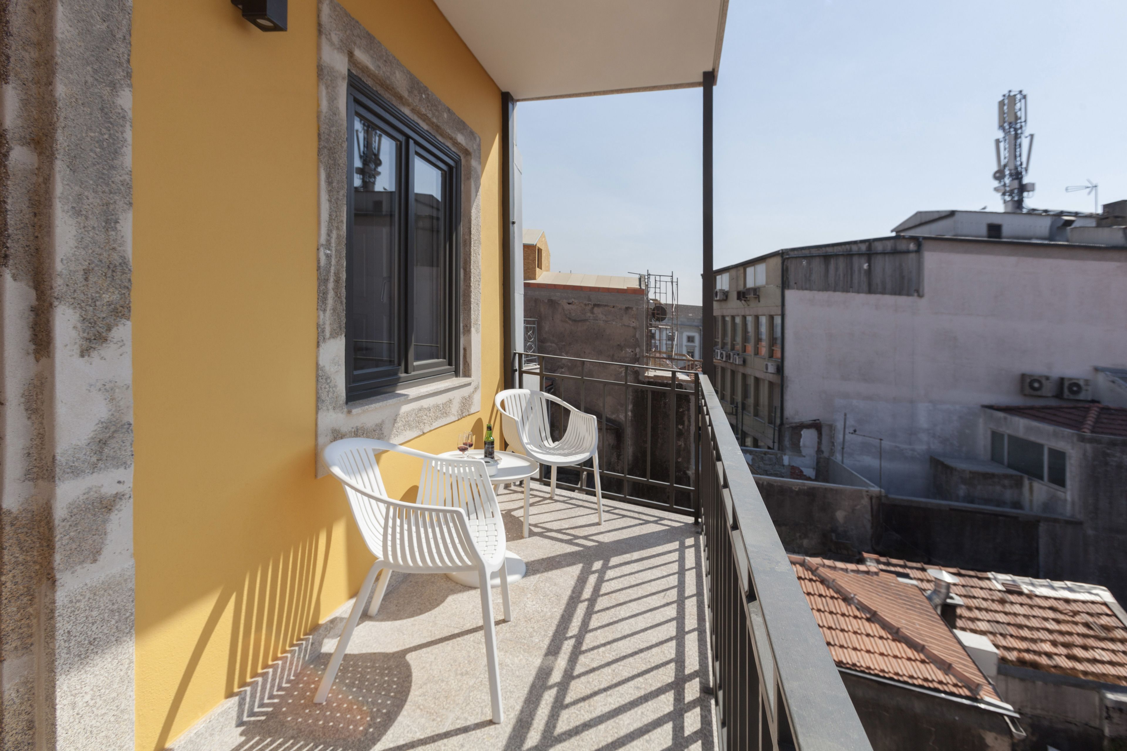 A balcony with white chairs and a small table, overlooking a cityscape with buildings and a clear sky.