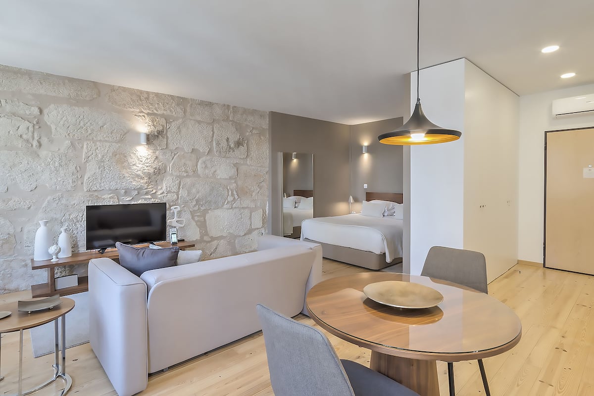 A modern hotel room with a stone accent wall, a flat-screen TV, a sofa, a dining table, and a bed.