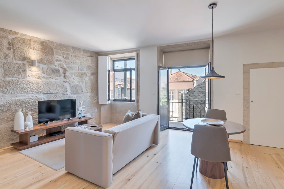 A modern living room with a stone wall, a flat-screen TV, a sofa, a dining table with chairs, and a balcony with a view.