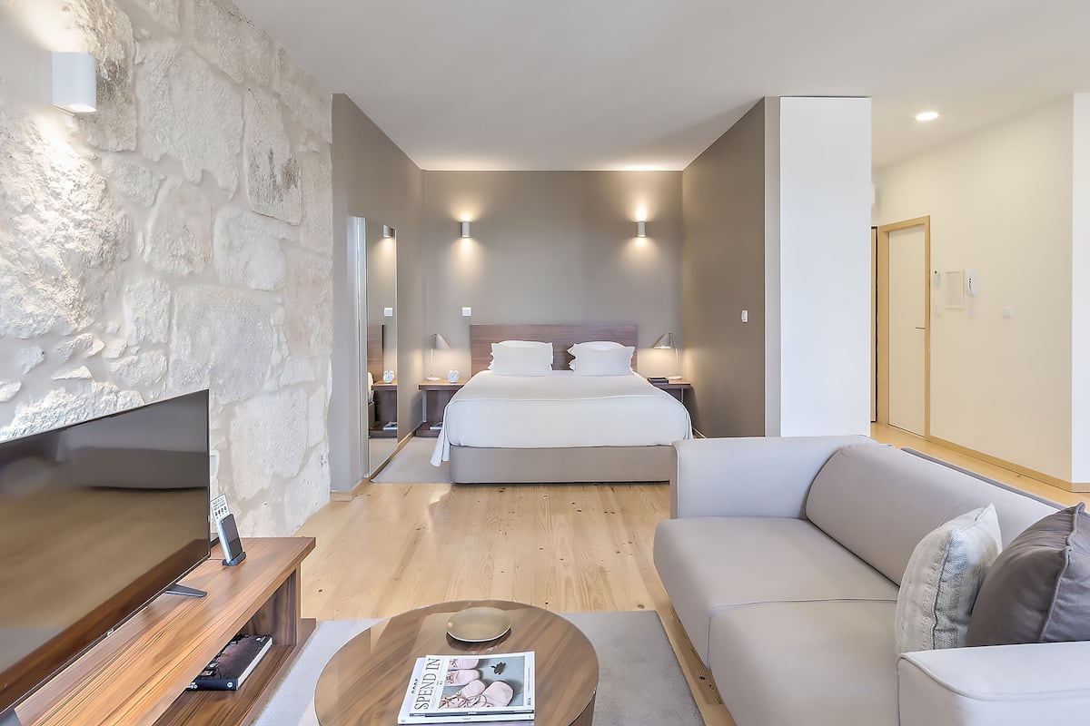 A modern hotel room with a stone accent wall, a large bed, a sofa, a TV, and a bedside table.