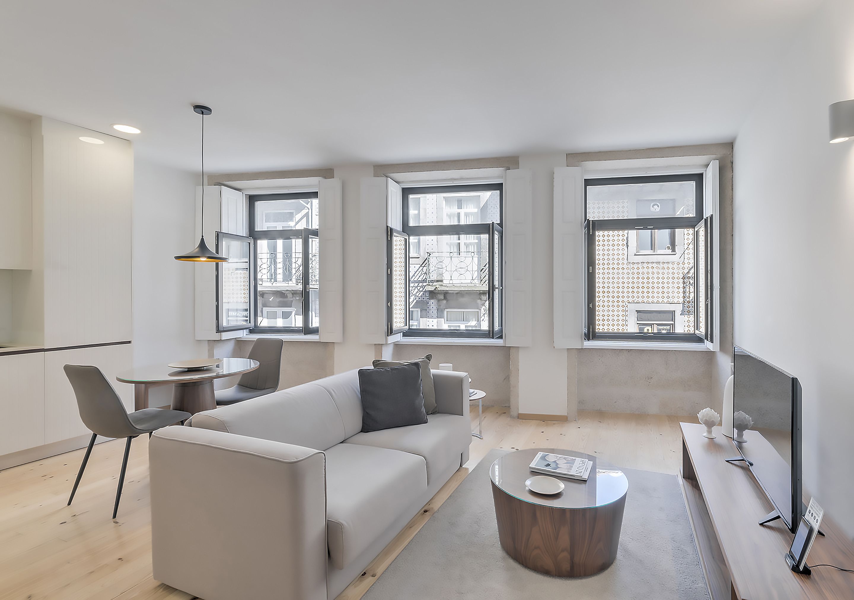 A modern living room with a sofa, coffee table, and dining area. There are large windows providing natural light and views of the outside.