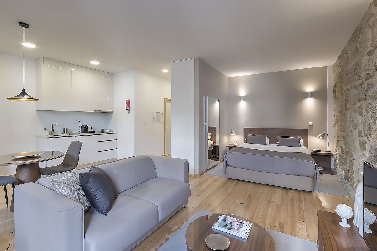 A modern hotel room with a sofa, a dining table, a kitchen area, and a separate bedroom with a bed and bedside table.