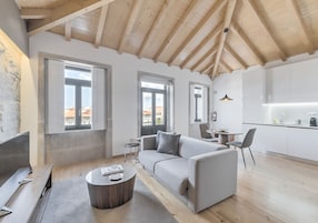 Uma sala de estar moderna com um sofá, mesa de centro e televisão. Janelas amplas e uma área de cozinha são visíveis.