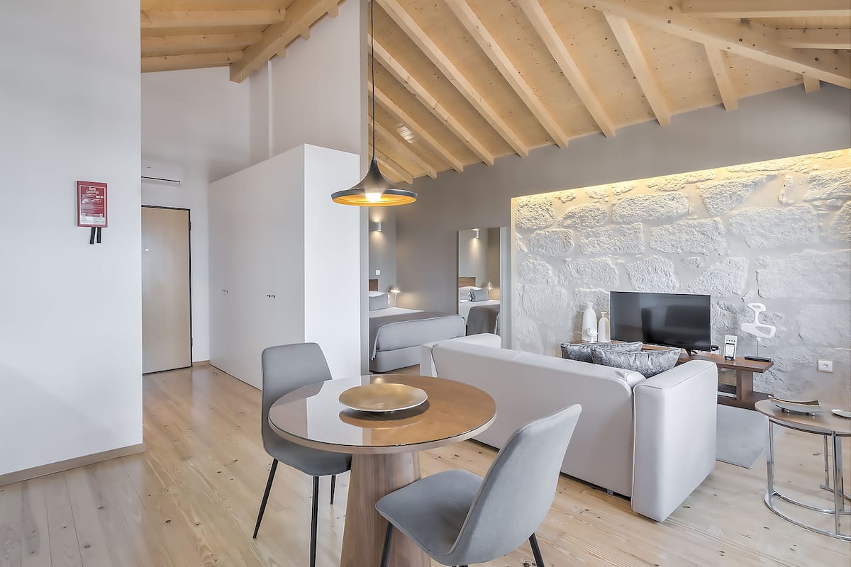 A modern living room with a wooden ceiling, a dining area with a round table and chairs, a sofa, and a TV on a stone wall.