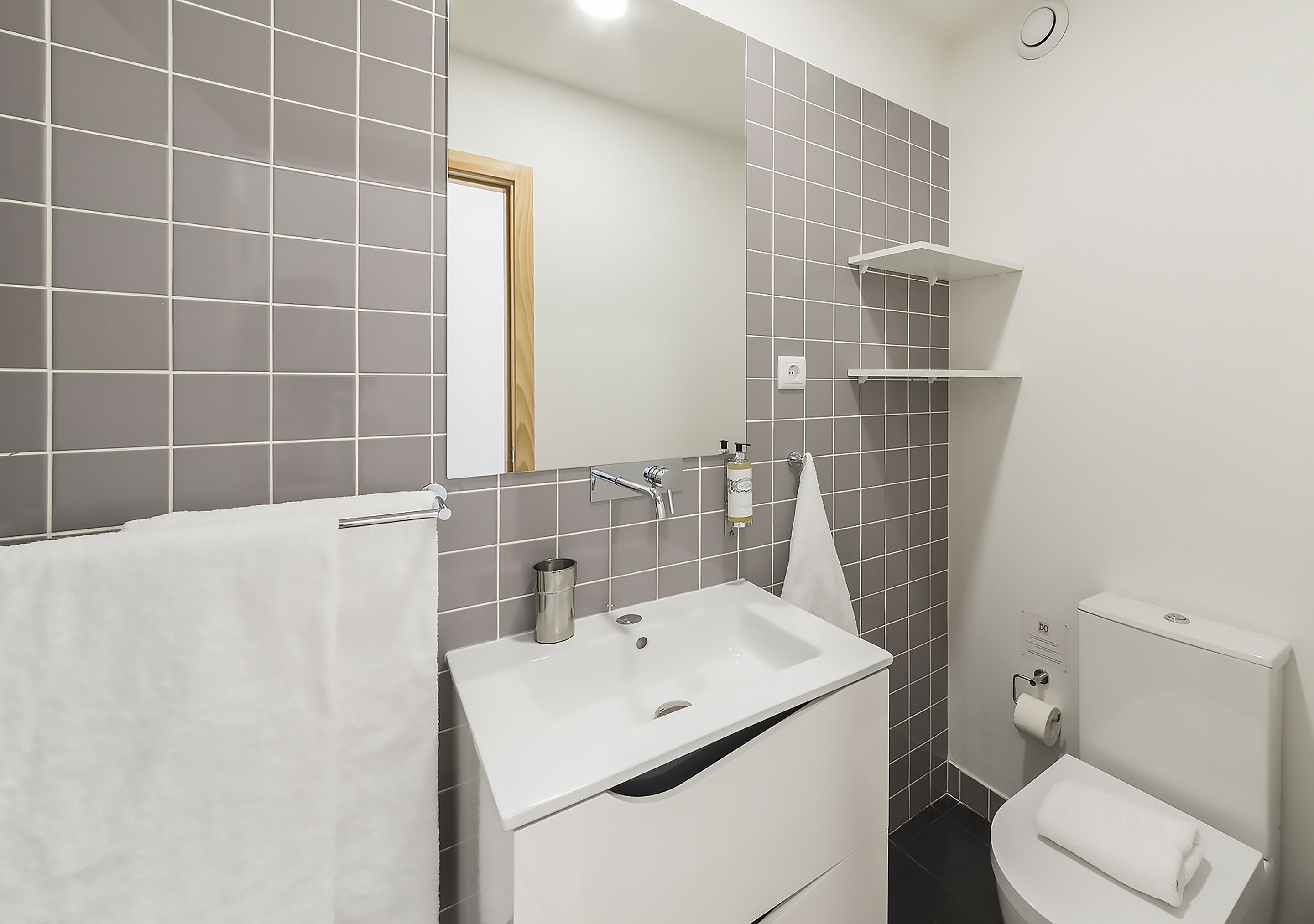 A modern bathroom with a white sink, mirror, and toilet, grey tiled walls, and a small white shelf.