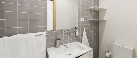 A modern bathroom with a white sink, mirror, and toilet, grey tiled walls, and a small white shelf.