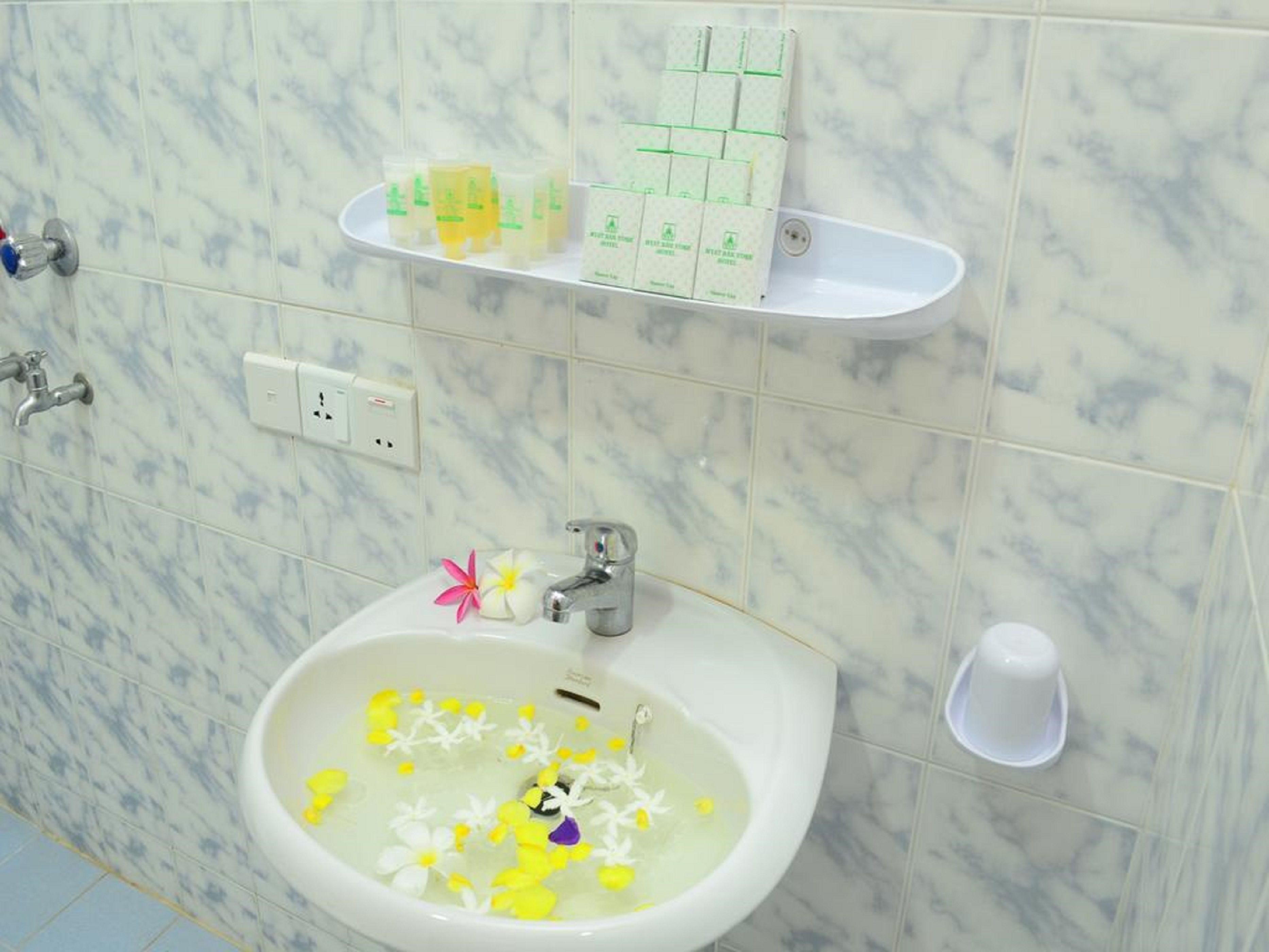 A bathroom with a marble-tiled wall, a sink with flowers, and toiletries on a shelf.