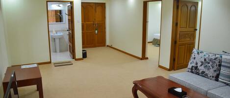 Amplia sala de estar con una mesa de centro de madera, un sofá con cojines estampados y una mesita con dispositivos electrónicos. Dos puertas de madera dan acceso a otras habitaciones, y el suelo está alfombrado.