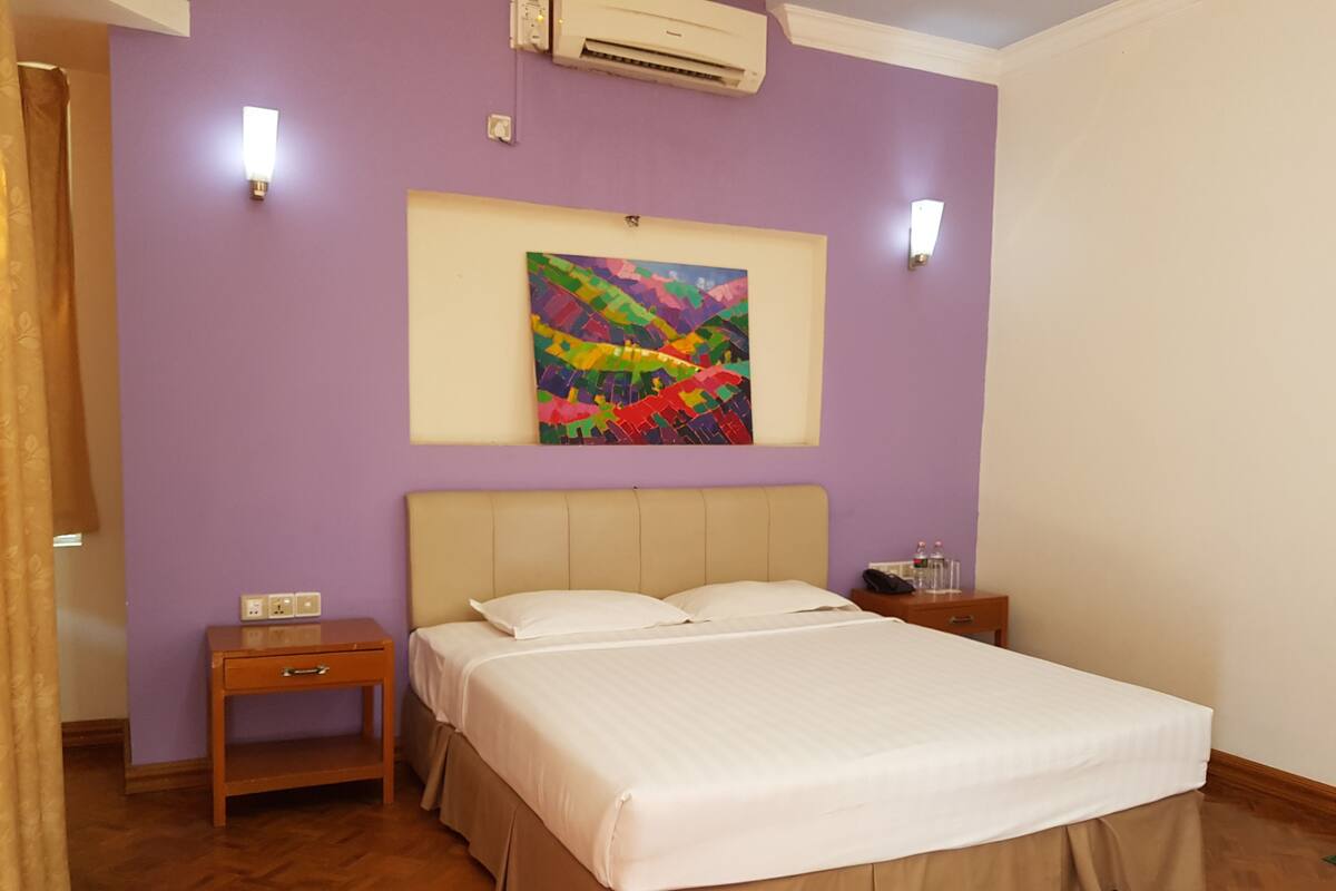 Une chambre d’hôtel avec un lit, une table de chevet, un climatiseur fixé au mur et un tableau abstrait aux couleurs vives.