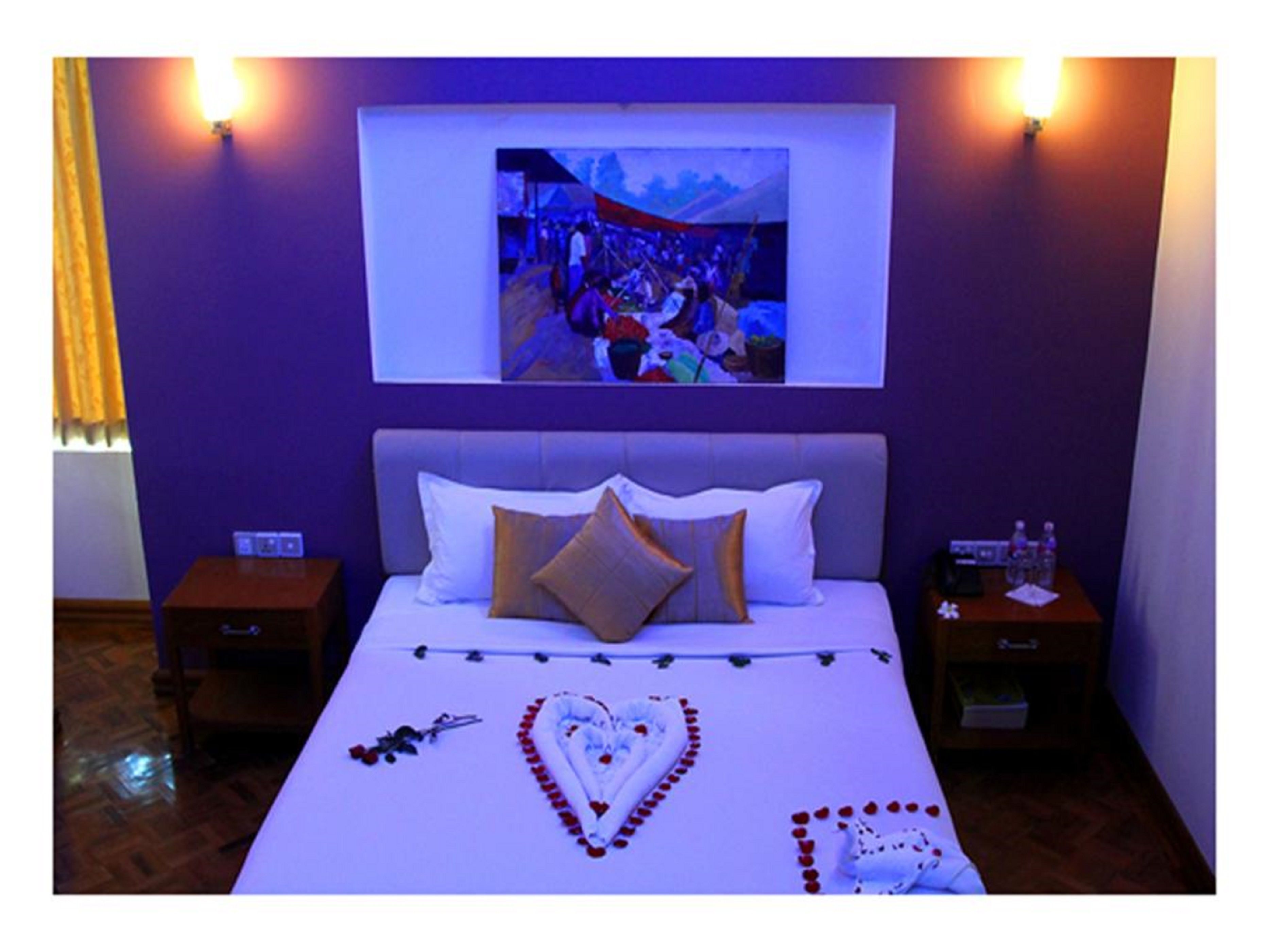 Hotelkamer met een bed, nachtkastje, tv aan de muur, en een decoratief hart van rozenblaadjes op het bed.