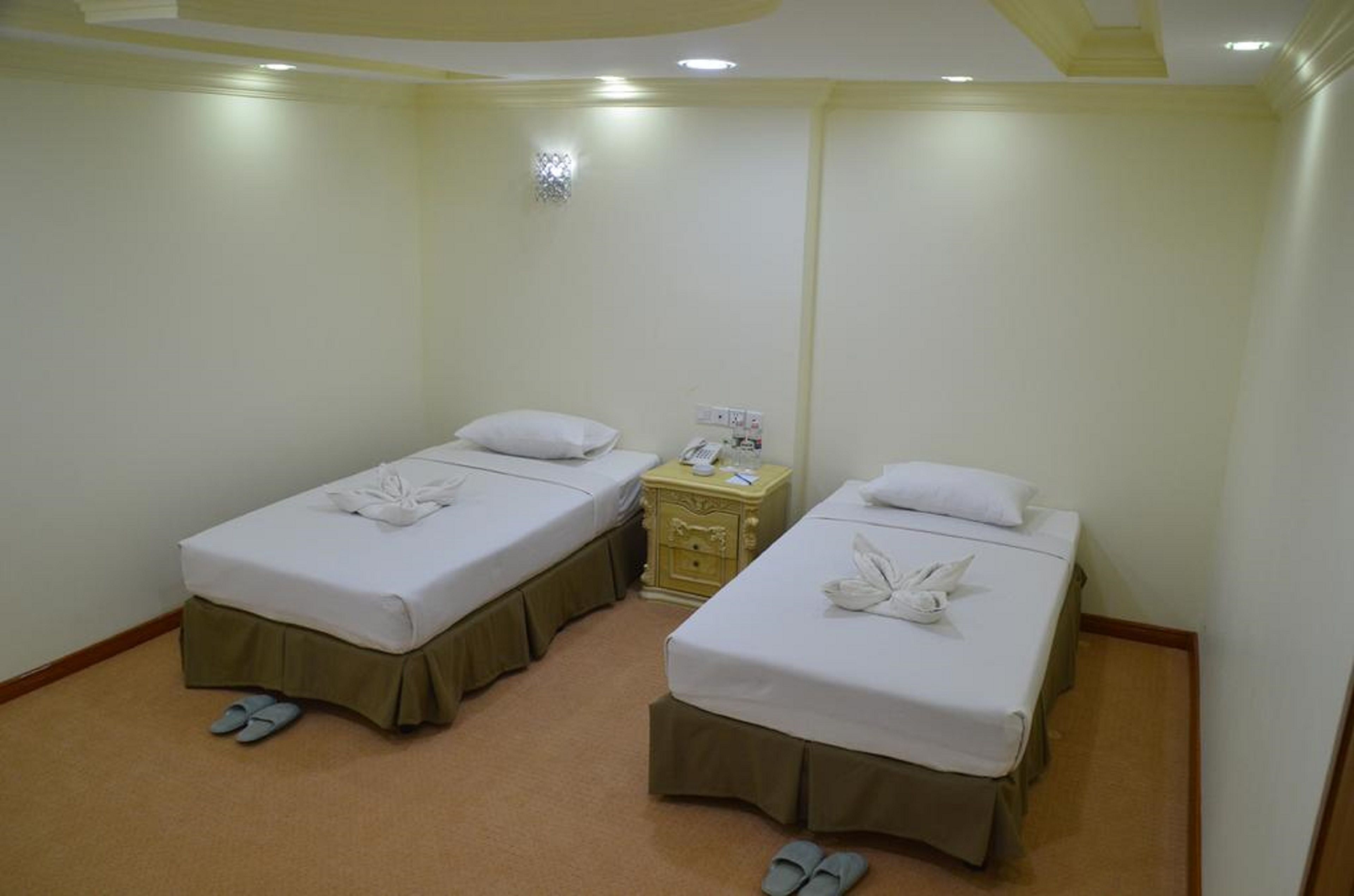 A hotel room with two single beds, each with a pillow and a towel. There is a small bedside table with a phone and a lamp. The room has beige walls and a carpeted floor.