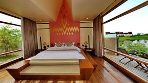 Una cama grande con estructura y cabecera de madera, con un diseño geométrico. Hay dos mesitas de noche con lámparas, una ventana con vistas a zonas verdes y un edificio, y una terraza de madera en el exterior.