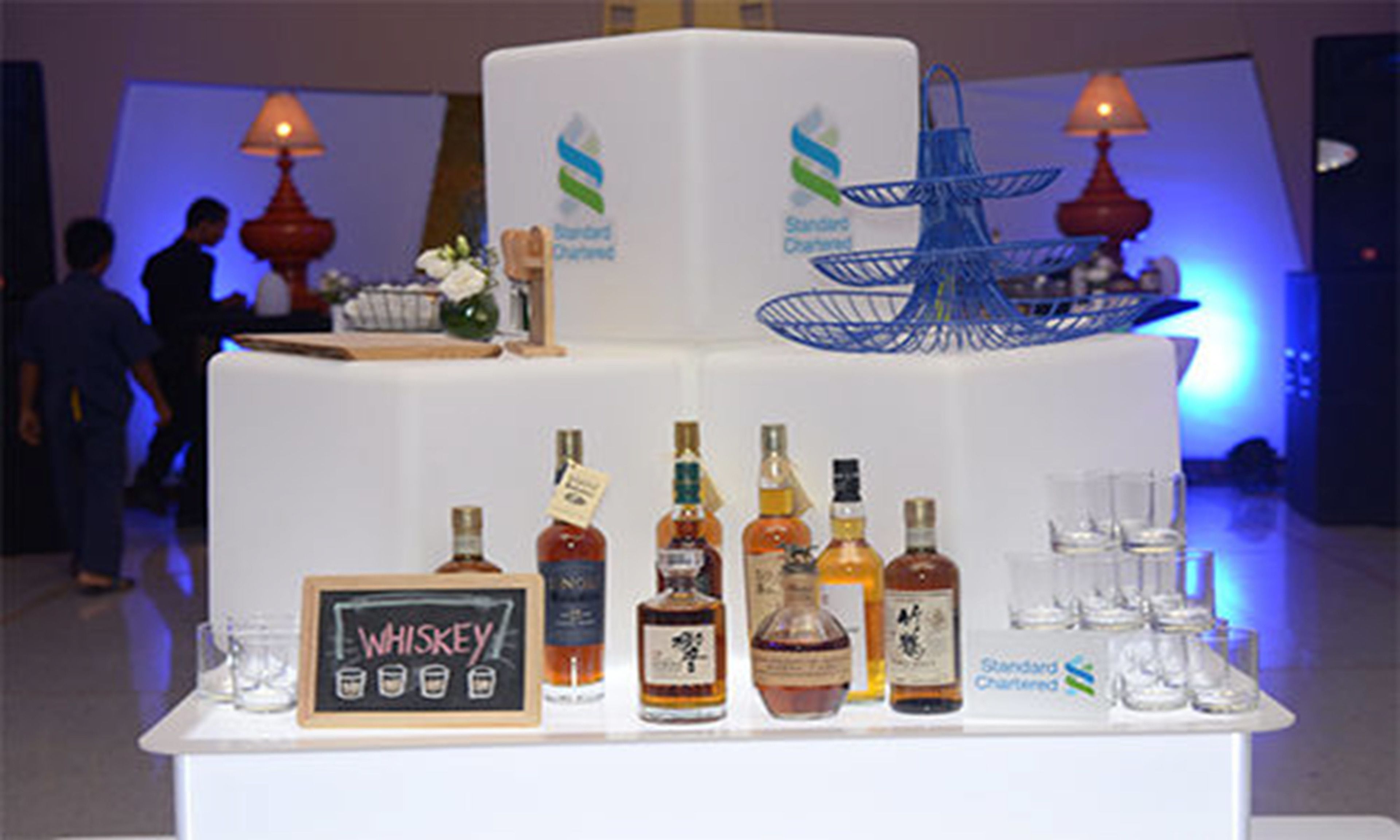 A multi-tiered bar setup with various liquor bottles and glasses, a chalkboard sign labeled "WHISKEY," and a blue decorative stand.