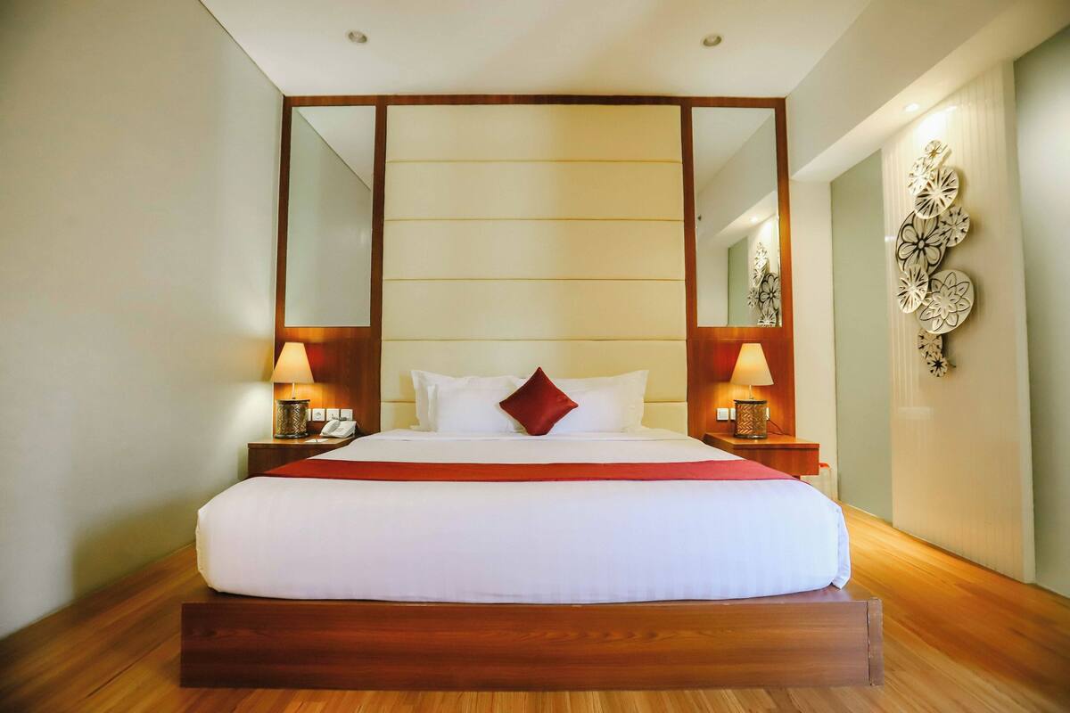 A hotel room with a large bed, wooden headboard, two bedside tables with lamps, a mirror, and decorative wall art.