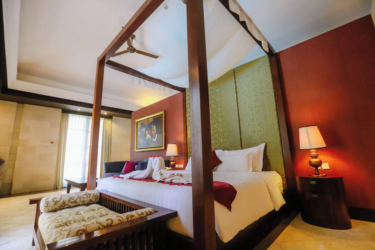 A four-poster bed with a canopy, a bedside table with a lamp, a chair, and a painting on the wall.