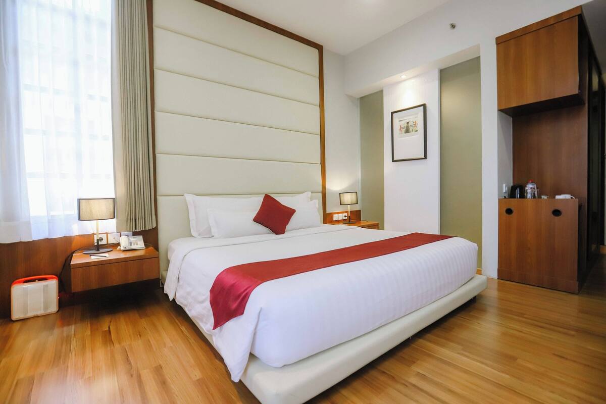 A hotel room with a large bed, wooden floor, bedside tables with lamps, a wardrobe, and a window with curtains.