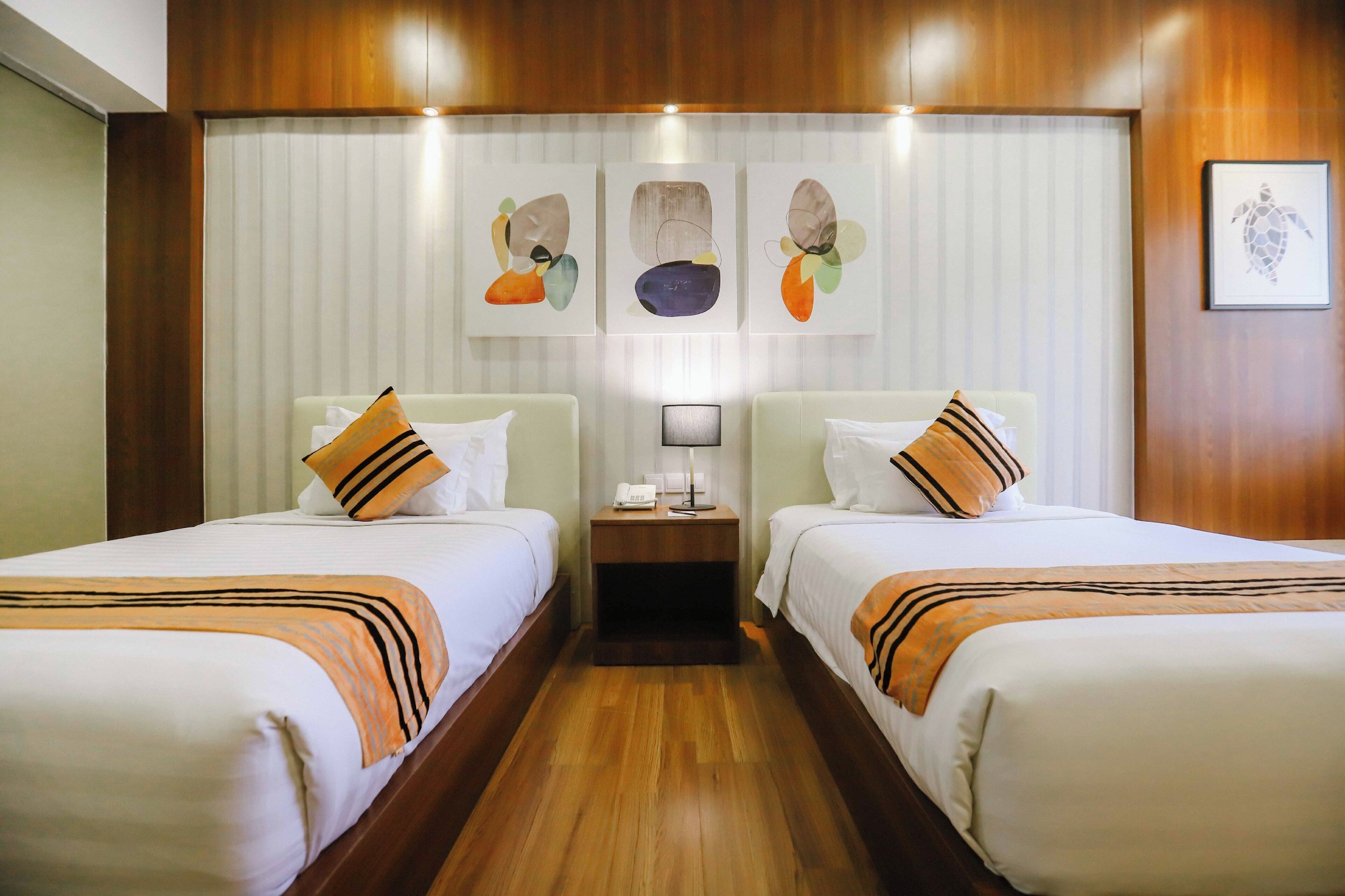 A hotel room with two beds, wooden flooring, a nightstand with a lamp, and artwork on the wall.
