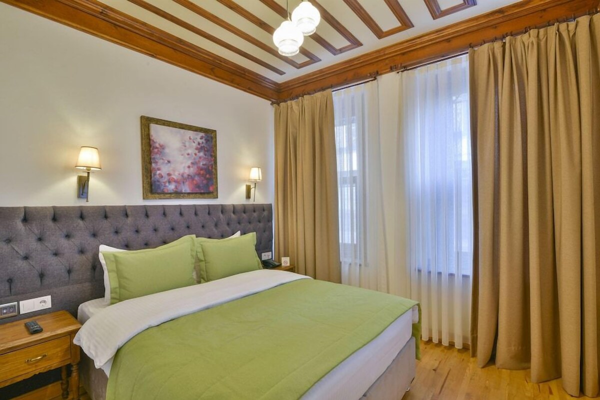 A hotel room with a tufted headboard, a green bedspread, two bedside lamps, a painting on the wall, and a large window with curtains.