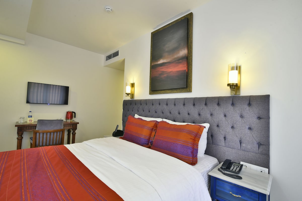 A hotel room with a bed, a desk, a chair, a TV, and a painting on the wall.